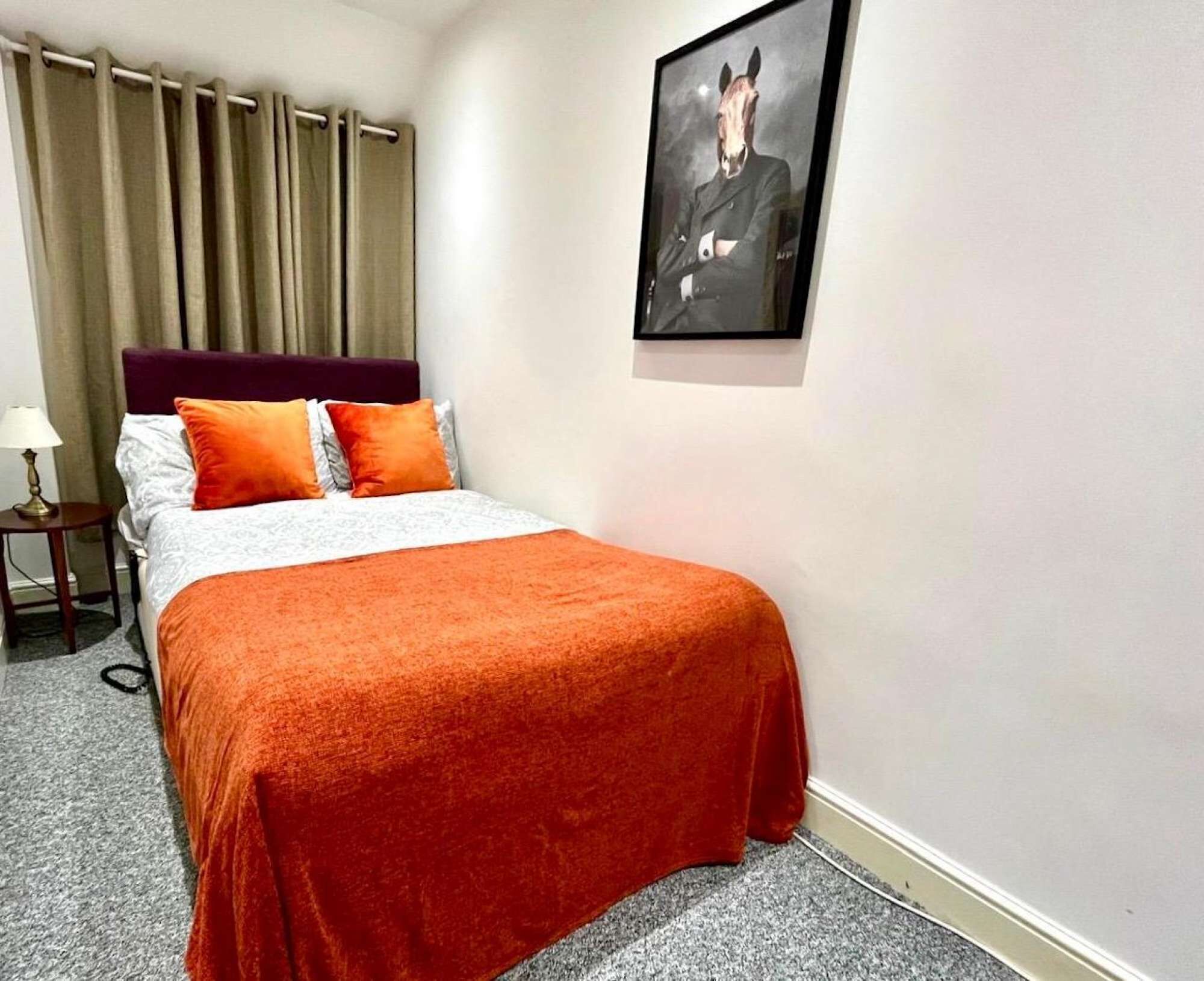 Habitación de hotel con cama, mesita de noche, lámpara, un cuadro enmarcado en la pared y ventana con cortinas.