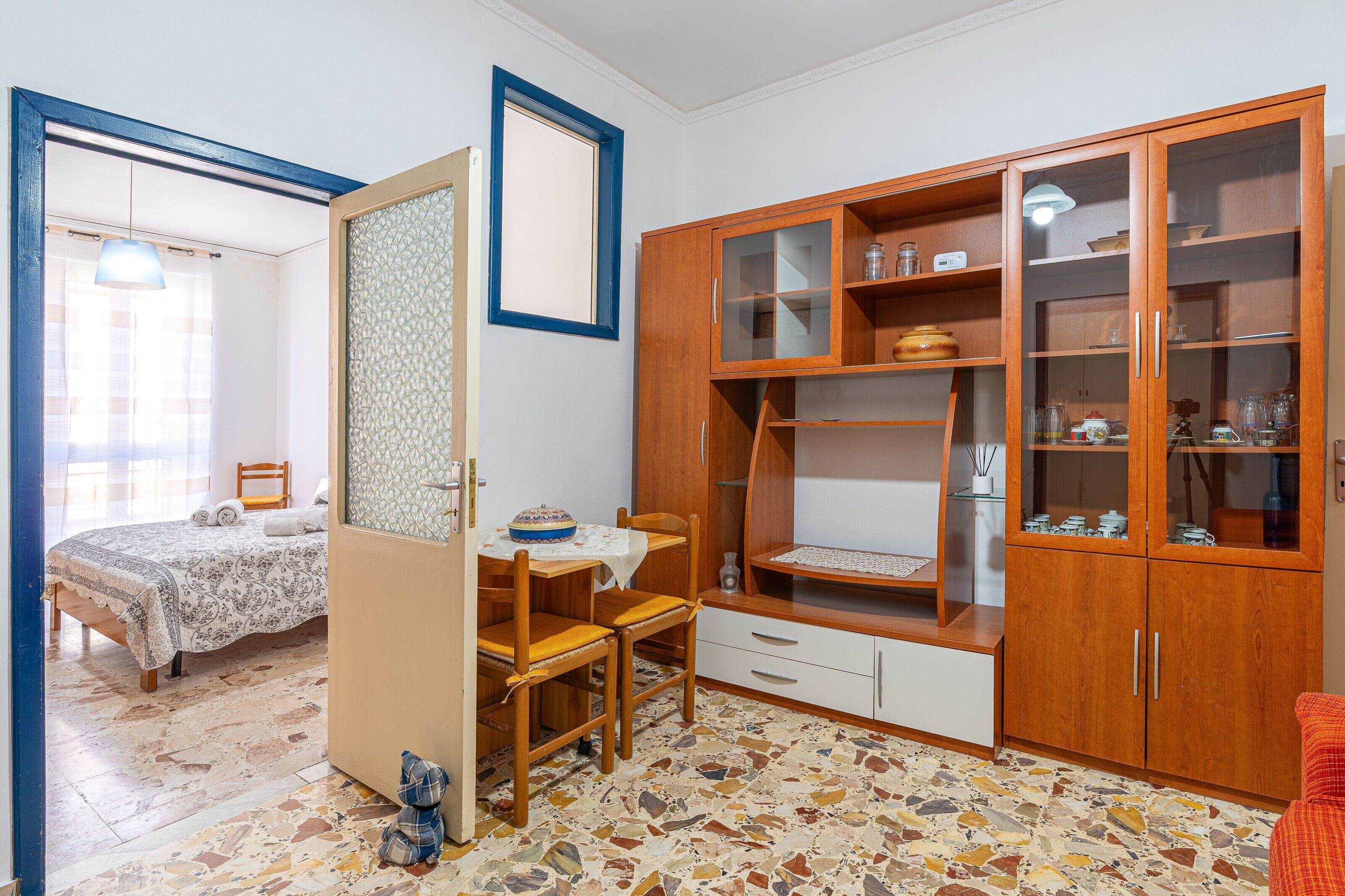 Ein Schlafzimmer mit Bett, einem Holzschrank mit Glastüren, einem Esstisch mit Stühlen und einem kleinen Tisch mit einem dekorativen Gegenstand.