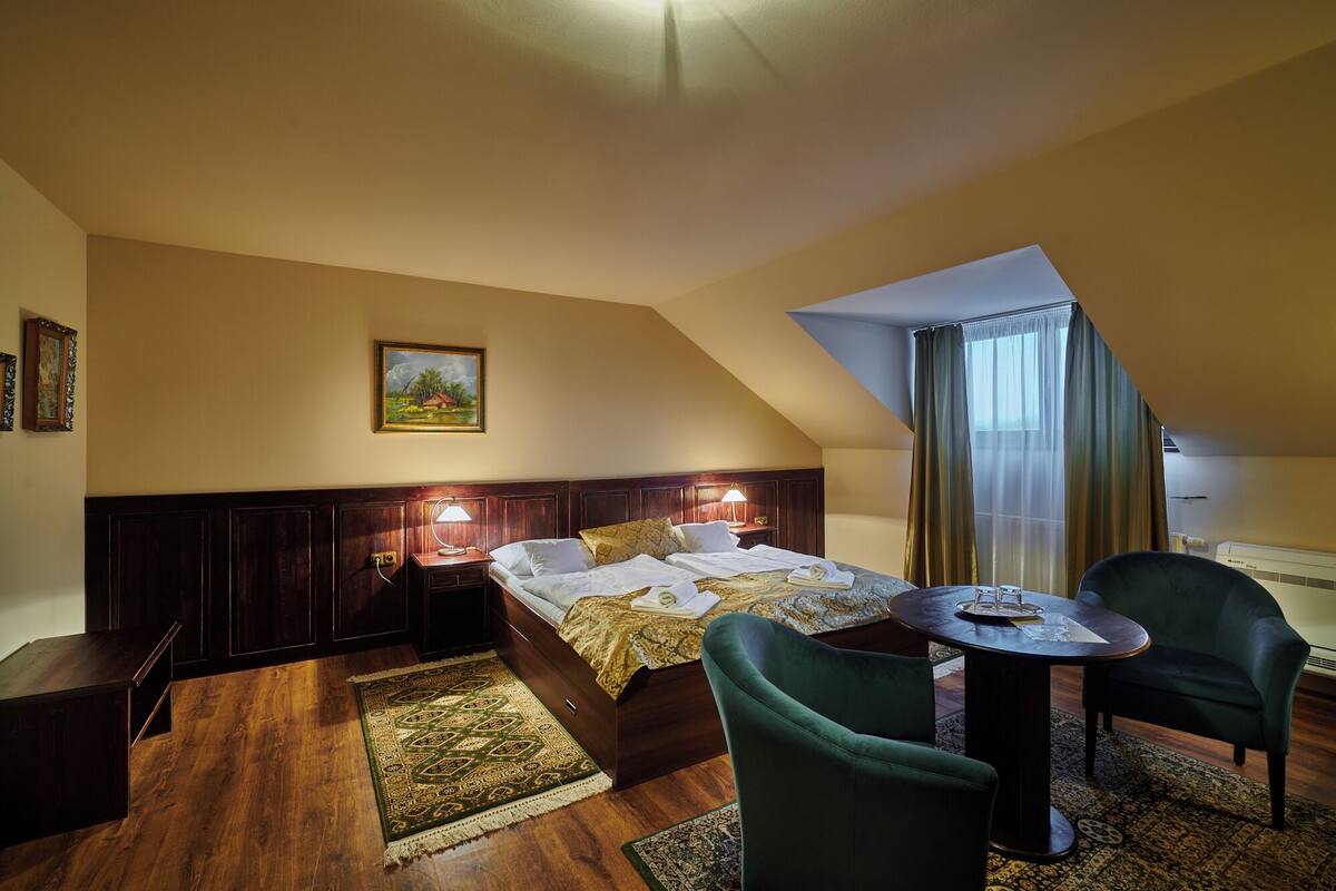 Ett hotellrum med en säng, två stolar, ett litet bord, en tavla på väggen och ett fönster med gardiner.