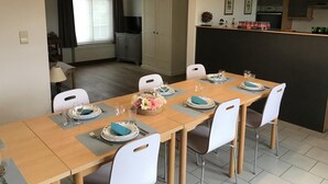 A dining area with a long table set for four, white chairs, a kitchen area with cabinets and a countertop, and a window with curtains. - Countryside Stay With Terrace (Heers)