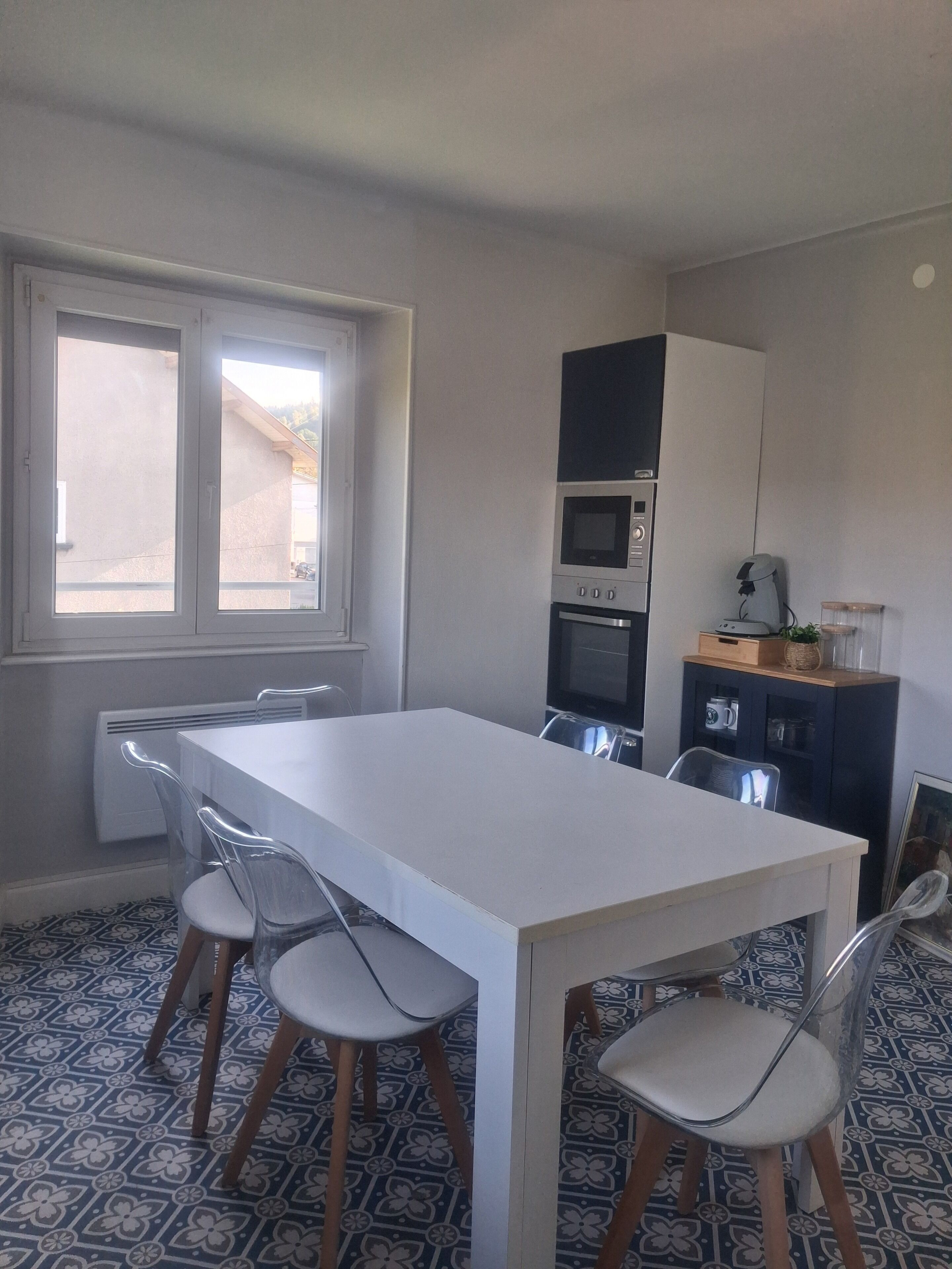 Uma sala de jantar com uma mesa e cadeiras brancas, uma cozinha com eletrodomésticos e uma janela com vista.