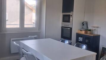 Uma sala de jantar com uma mesa e cadeiras brancas, uma cozinha com eletrodomésticos e uma janela com vista.