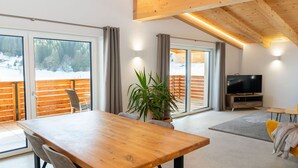 A modern living room with a wooden dining table, a sofa, a TV, and large windows with a view of a snowy landscape. - Experience beautiful high-ceilinged living rooms with exposed roof trusses! (Wildschönau)
