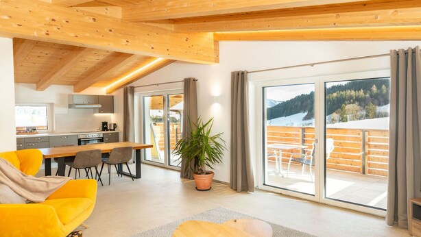 A modern living room with a sofa, dining table, and chairs, a kitchen area, and a balcony with a view of the mountains. - Experience beautiful high-ceilinged living rooms with exposed roof trusses! (Wildschönau)