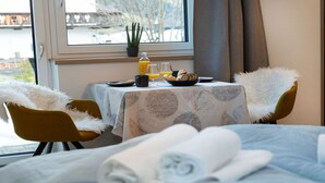 Une chambre d’hôtel avec une table à manger dressée pour le petit-déjeuner, un lit avec des serviettes pliées et une fenêtre donnant sur un pont et des arbres.
