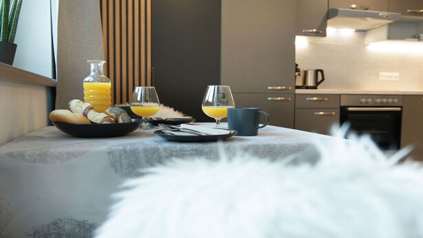 Une table de petit-déjeuner avec un bol de pain, un verre de jus d’orange et une tasse, le tout disposé sur une table dans une cuisine moderne équipée d’une cuisinière et de placards.