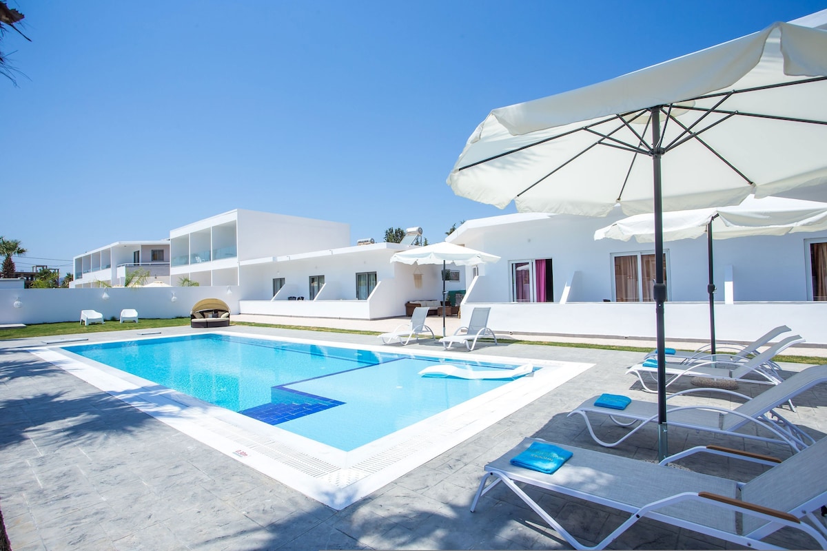 Ein Swimmingpool mit Liegestühlen und Sonnenschirmen, umgeben von weißen Gebäuden.
