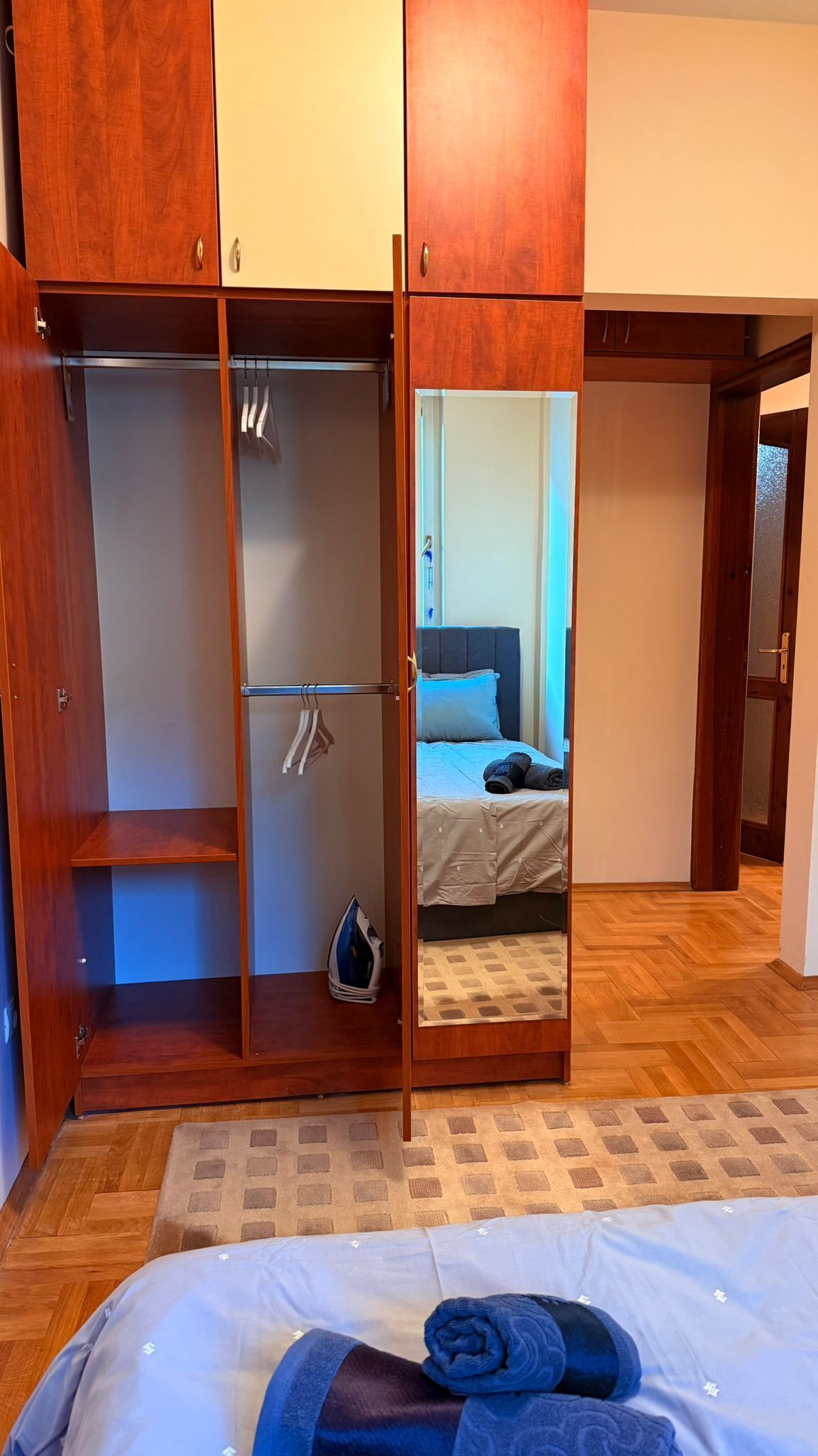 Ein Schlafzimmer mit einem Holzschrank, einem Bett mit blauer Kopfteil und einem Spiegel, der den Raum widerspiegelt.