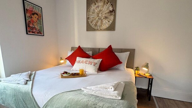 Een net opgemaakt bed met een witte deken, rode en witte kussens, en een kussen met de tekst 'BEACH'. Er liggen handdoeken op het bed en een dienblad met fruit en sap. Een lamp en een klein tafeltje met een plant staan ernaast.