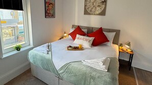 Een netjes opgemaakt bed met een schaal fruit en een fles water, een raam met uitzicht op gebouwen en een kleine plant op de vensterbank.
