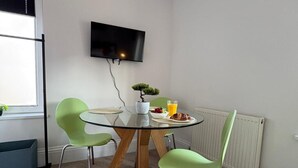 Een eetruimte met een ronde glazen tafel, twee groene stoelen, een kleine plant in pot, een aan de muur bevestigde televisie en een radiator.