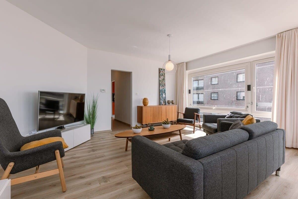 A modern living room with a flat-screen TV, grey sofas, a wooden coffee table, and a large window with curtains.