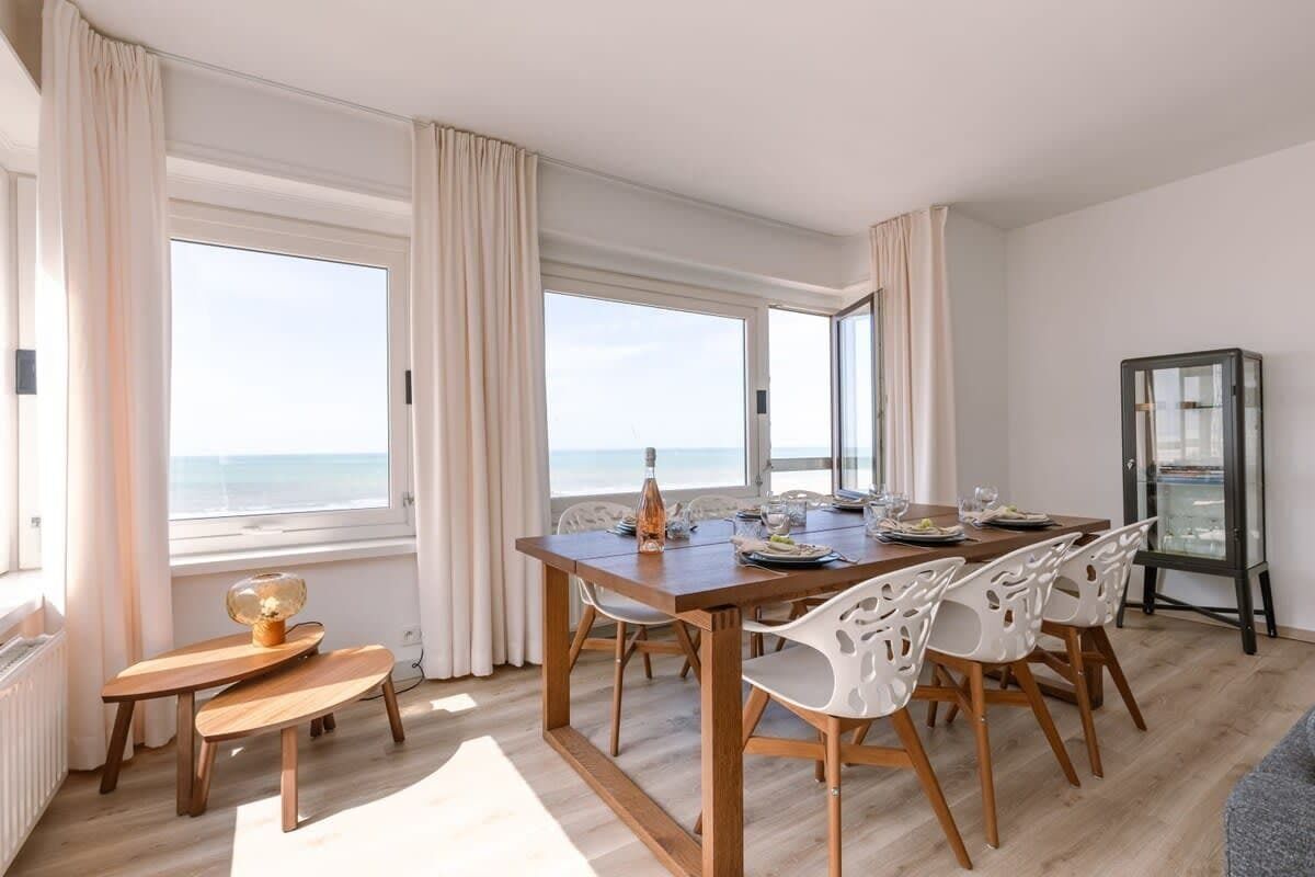 A dining area with a wooden table, white chairs, and a view of the sea through large windows. - Waterfront retreat w/ Wifi & workspace (Middelkerke)