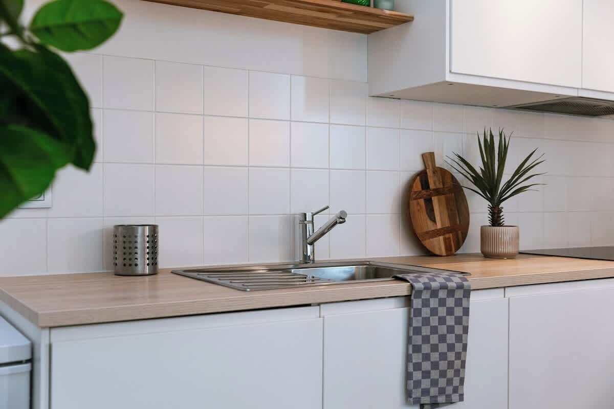Una cocina con armarios blancos, un lavamanos de acero inoxidable, una tabla de cortar de madera y una planta en maceta.