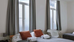 Ein Hotelzimmer mit zwei Betten, grauen Vorhängen, einem weißen Stuhl und einem Fenster.