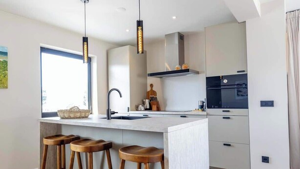 A modern kitchen with a central island, bar stools, pendant lighting, and a large window. - Coastal apartment w/ sauna & balcony (Cadzand)