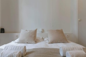 Een net opgemaakt bed met opgerolde handdoeken, een nachtkastje met een lamp en een aan de muur bevestigde thermostaat.
