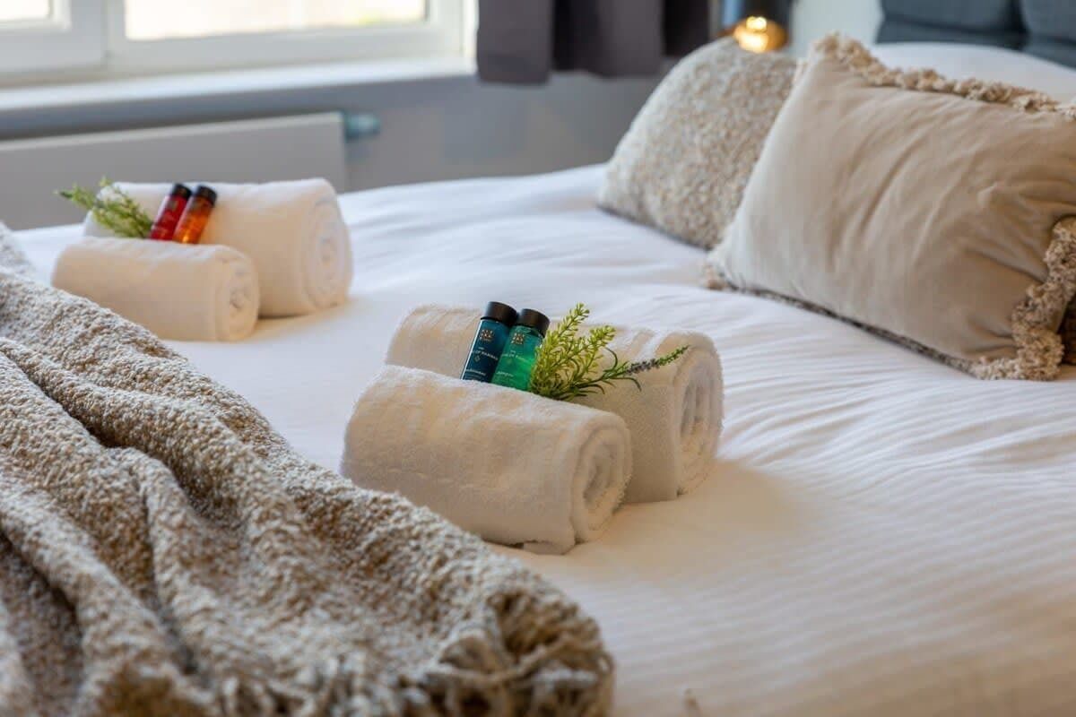 Rolled towels with toiletries arranged on a neatly made bed with textured pillows and a cozy throw.