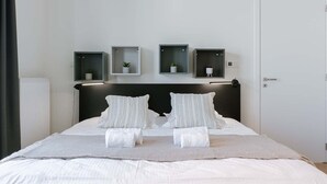 Une chambre d’hôtel moderne avec un grand lit, du linge de lit blanc, deux tables de chevet avec des lampes et trois étagères murales ornées de plantes.
