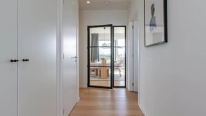 Un couloir aux murs blancs, doté d’un plancher en bois, une porte en verre donnant sur un coin repas, et un tableau encadré accroché au mur.