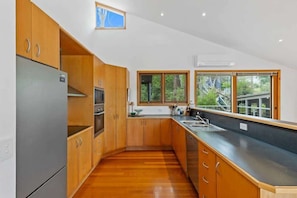 A modern kitchen with wooden cabinets, a stainless steel refrigerator, a built-in oven, and a large window providing a view of greenery. - Panorama at Rosedale (Rosedale)