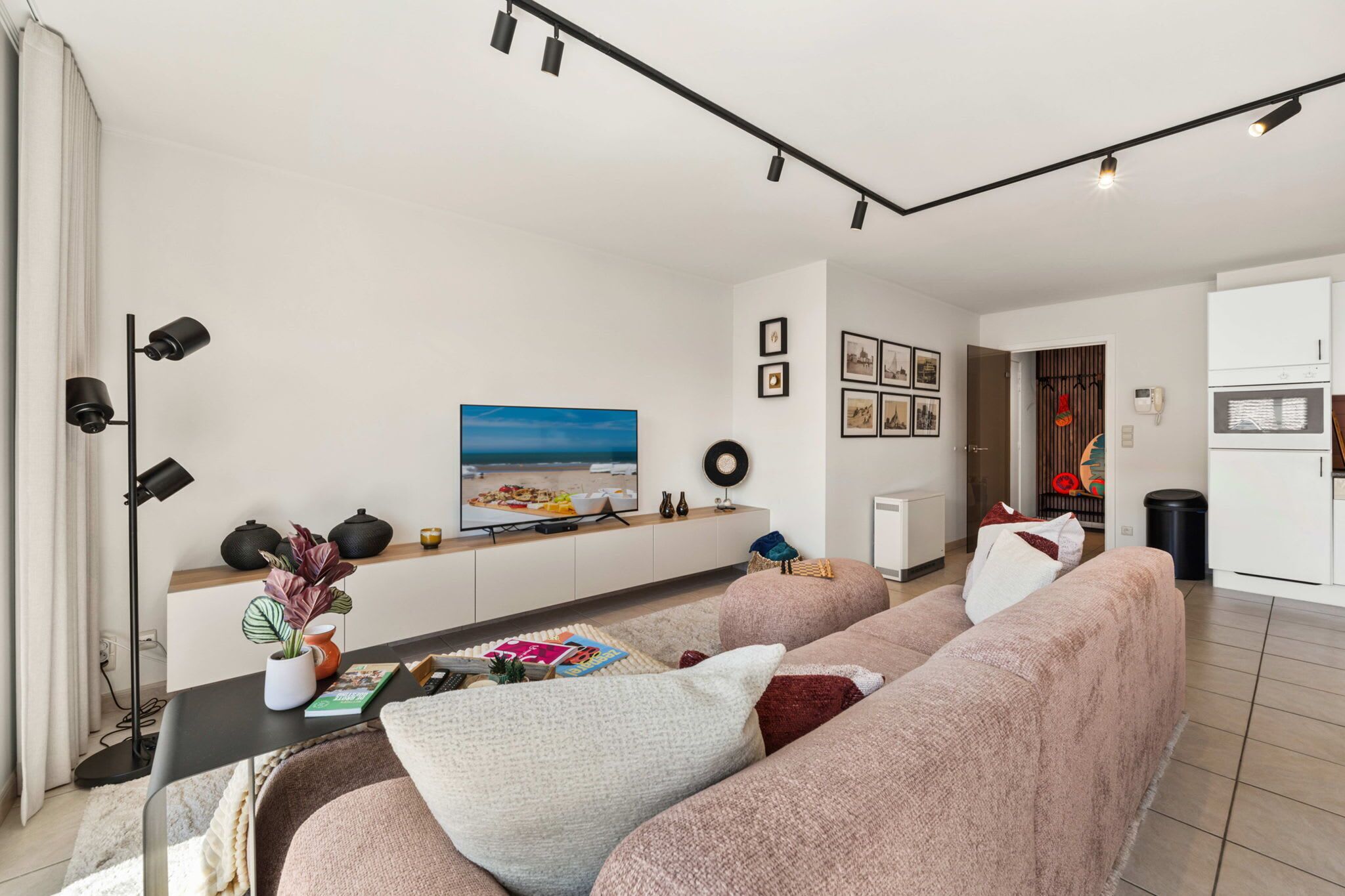 Una sala moderna con sofá, mesa de centro y televisor de pantalla plana. Se observa una zona de cocina con refrigerador y microondas.