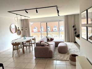 A modern living room with a sofa, dining table, and chairs, large windows with a view of red-roofed buildings, and a ceiling track lighting system. - Coastal retreat near Beach at Koksijde (Koksijde)