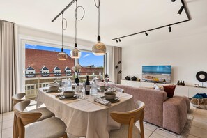 A dining area with a table set for four, a sofa, and a television in the background. - Beachfront Patio Apt in Koksijde w/ Natural Light (Koksijde)
