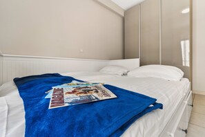 A bedroom with a bed, white bedding, a blue blanket, and a magazine on the bed. - Beachfront Patio Apt in Koksijde w/ Natural Light (Koksijde)