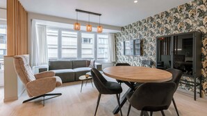 Ein modernes Wohnzimmer mit einer Couch, einem Esstisch mit Stühlen, einem an der Wand befestigten Fernseher und großen Fenstern.