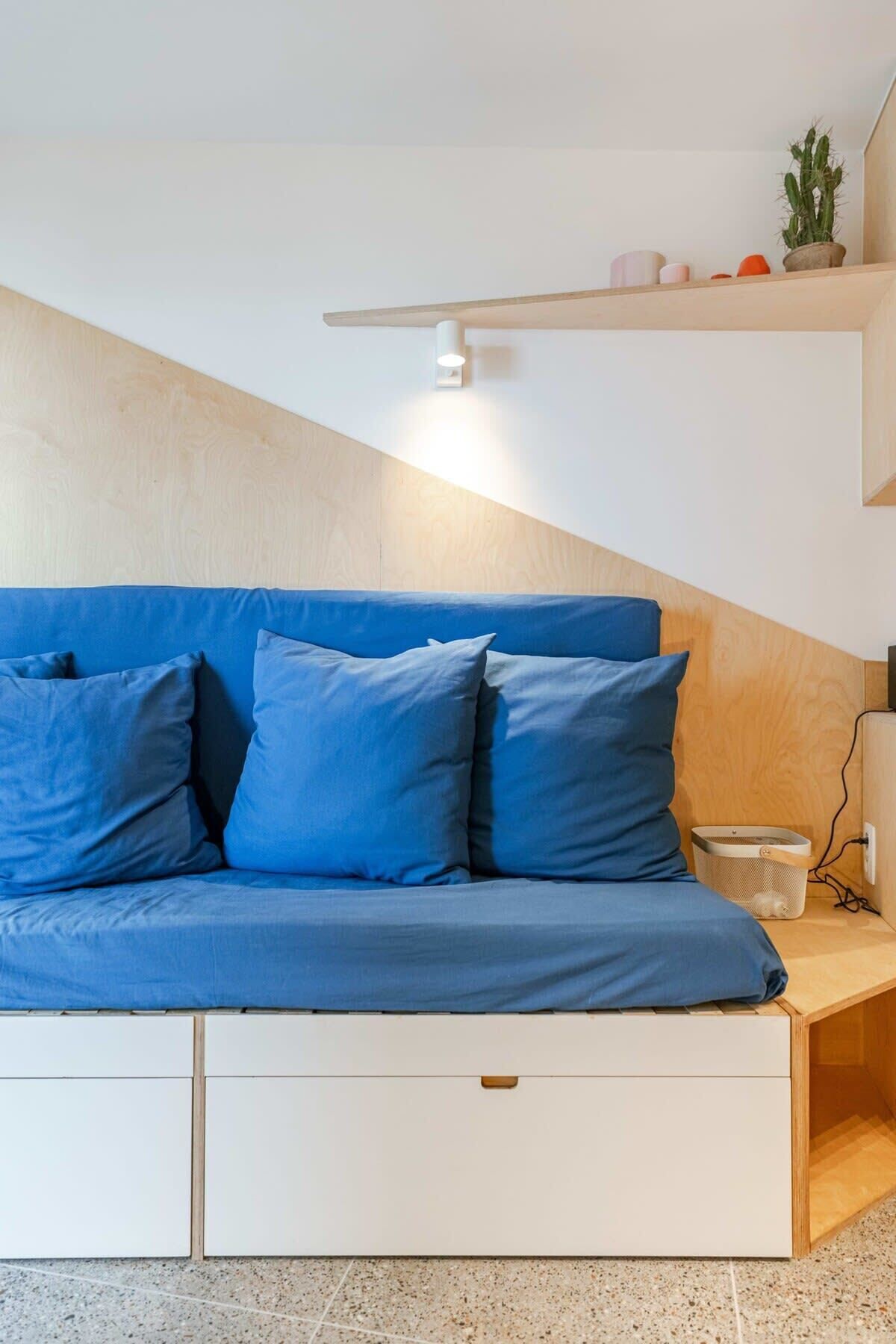 A modern bedroom with a blue sofa, a wooden headboard, a small shelf with decorative items, and a bedside table with a lamp.