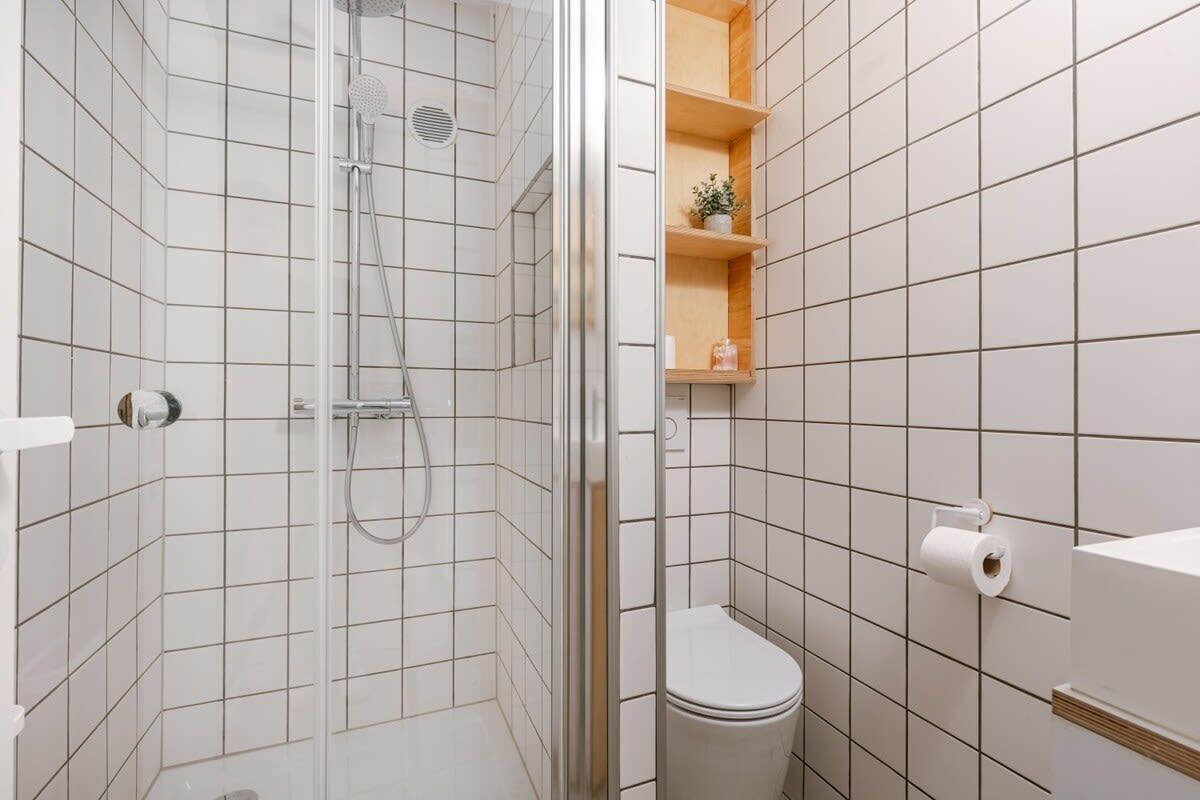 Ein Badezimmer mit weißen Fliesen, einer Glasdusche, einer Toilette und Holzregalen mit Pflegeprodukten.