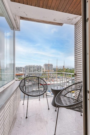 A balcony with two wicker chairs, a small table, and a view of a marina with boats and buildings. - Waterfront apartment w/ balcony & elevator (Oostende)