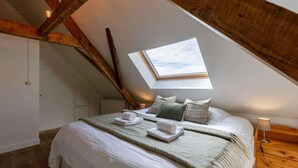 Un dormitorio con tragaluces, vigas de madera, una cama con ropa de cama blanca y una mesita de noche con lámpara.