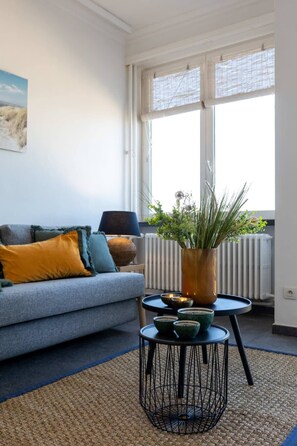 Ein Wohnzimmer mit grauem Sofa, einem kleinen Tisch mit Pflanzenvase und einer Heizung.