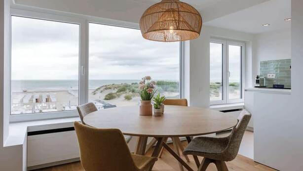 A modern dining area with a large window overlooking a beach, a round wooden table, and a woven pendant light. - Coastal getaway w/ full kitchen & workspace (Knokke-Heist)