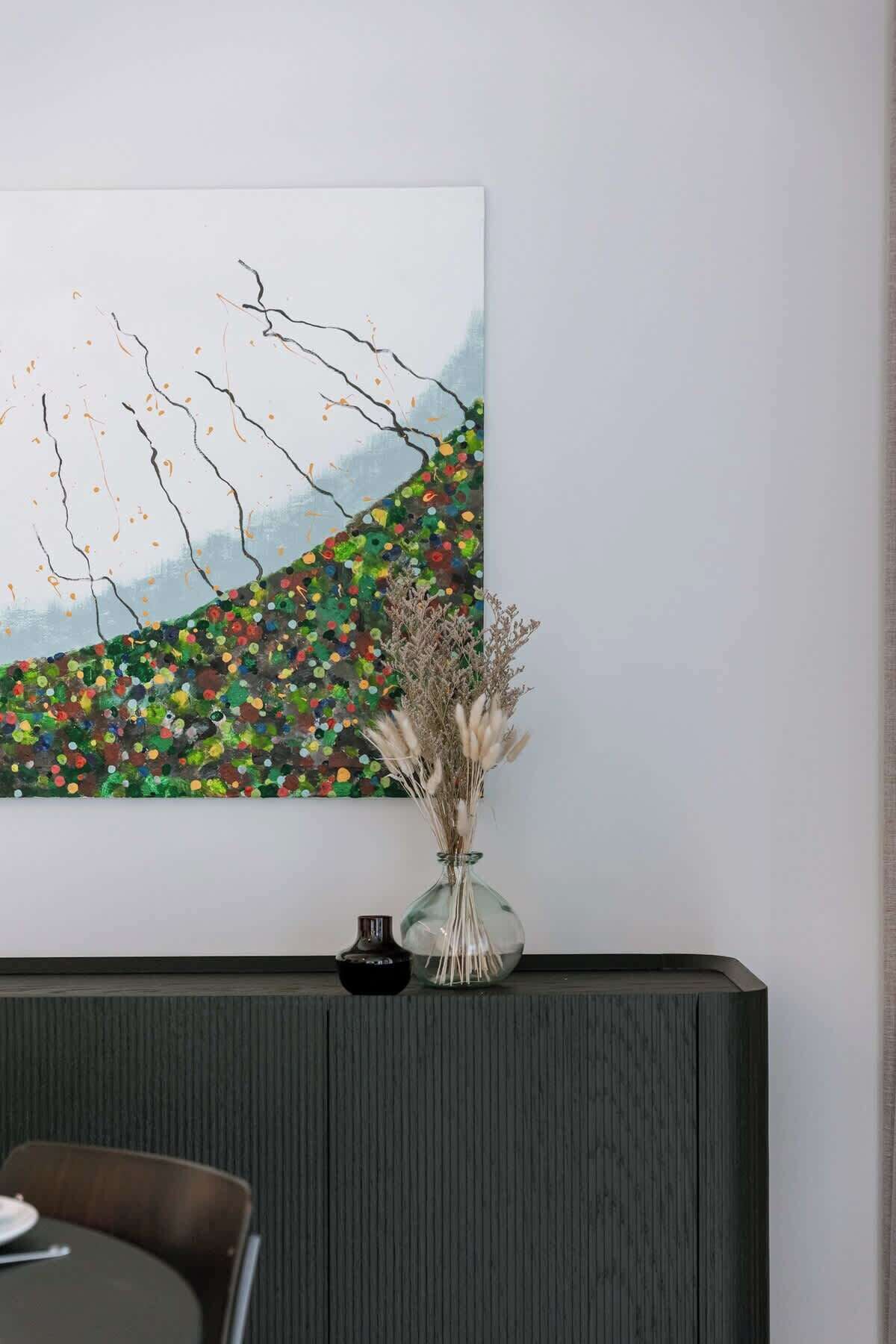 Un comedor con una mesa, sillas, un jarrón con plantas secas y un cuadro abstracto grande en la pared.