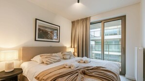 Ein Hotelzimmer mit Bett, Nachttisch inklusive Lampe, einem gerahmten Bild an der Wand und Blick auf ein Gebäude durch das Fenster.