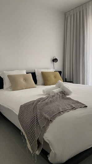 Ein ordentlich bezogenes Bett mit weißen und beigefarbenen Kissen, einer gemusterten Tagesdecke und einer schwarzen Wandlampe.