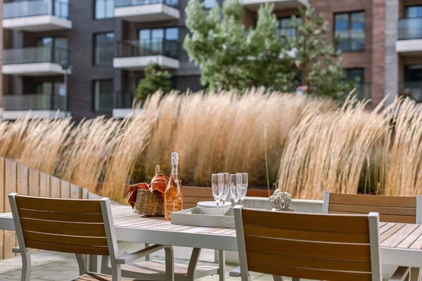Outdoor dining area with wooden table and chairs set against ornamental grasses and modern apartment balconies.