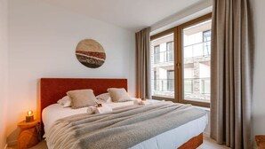 Et soveværelse med en stor seng, et sengebord med lampe, et vindue med gardiner og et kunstværk på væggen.