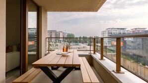 En balkon med et træbord og bænke, en glasdør samt udsigt til bygninger og byggeri i baggrunden.