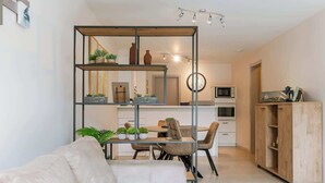 Un salon moderne comprenant un canapé, une table à manger avec des chaises, un coin cuisine avec des placards et des appareils électroménagers, et une bibliothèque en bois.