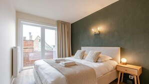 A bedroom with a large bed, a bedside table with a lamp, a view of the outside, and a radiator. - Waterfront retreat w/ beach access (De Panne)