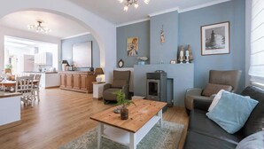 A spacious living area with a wooden coffee table, a sofa, and a fireplace. There is a kitchen in the background, a dining area with a table and chairs, and a large window providing natural light.