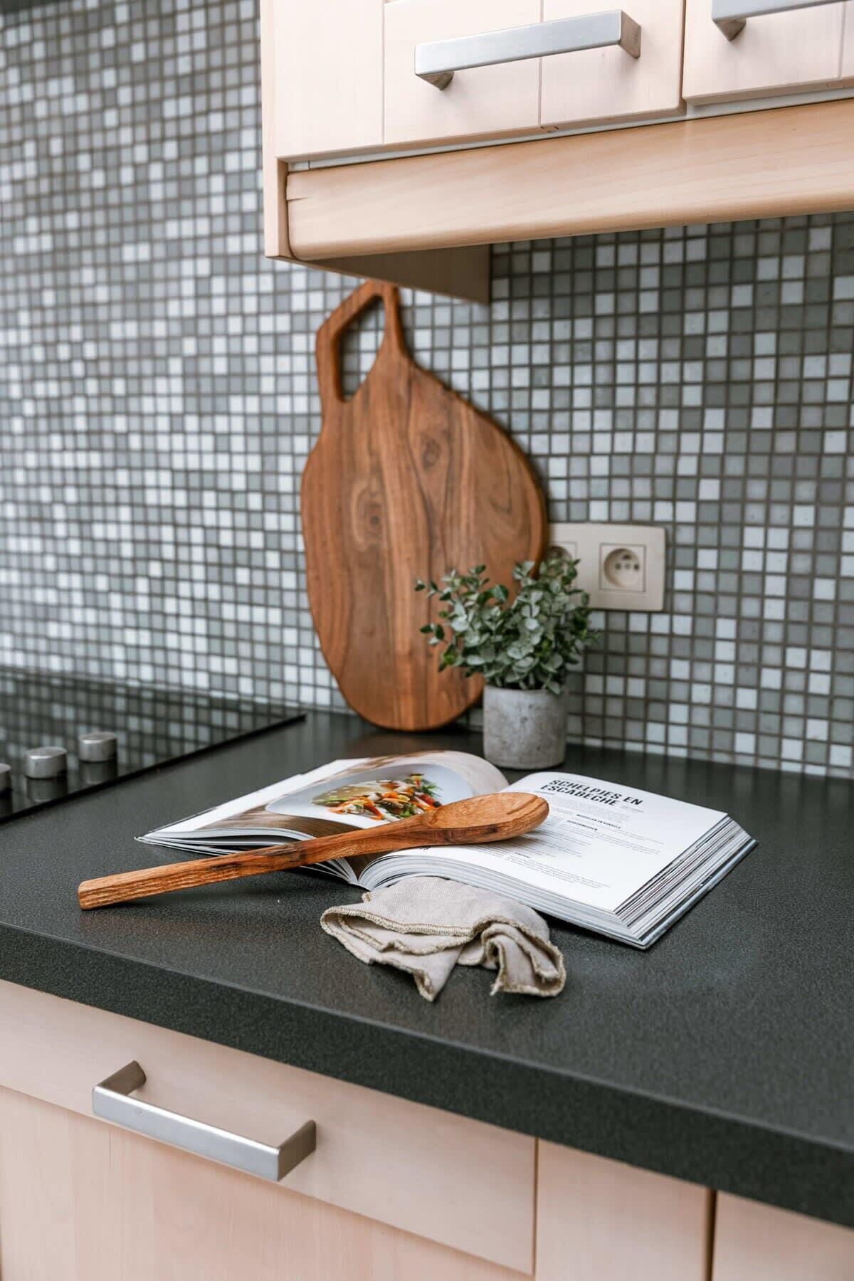 A kitchen counter with a wooden cutting board, a cookbook, a wooden spoon, and a cloth.