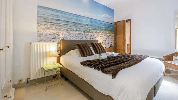 A bedroom with a large beach mural, a bed with a brown blanket, a bedside table with a lamp, and a wicker chair.