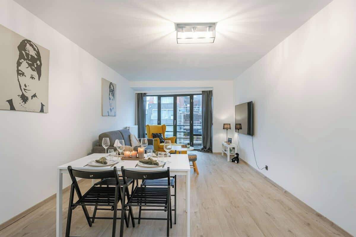 Un salon moderne avec une table à manger pour deux personnes, un canapé, un téléviseur et une grande fenêtre avec des rideaux.