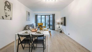 Ein modernes Wohnzimmer mit Esstisch für zwei Personen, einer Couch, einem Fernseher und einem großen Fenster mit Vorhängen.