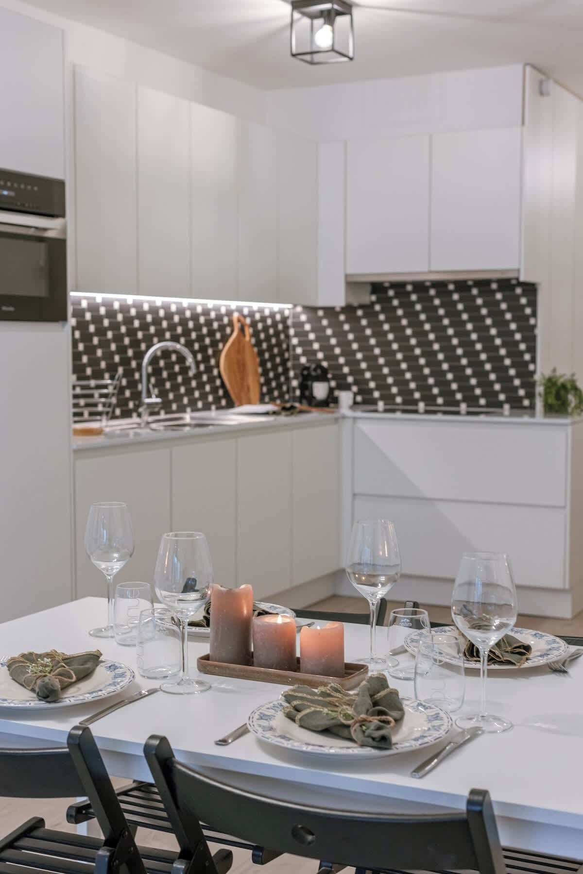 A modern dining area with a white table set for a meal, a kitchen with a black and white backsplash, and a ceiling light fixture.